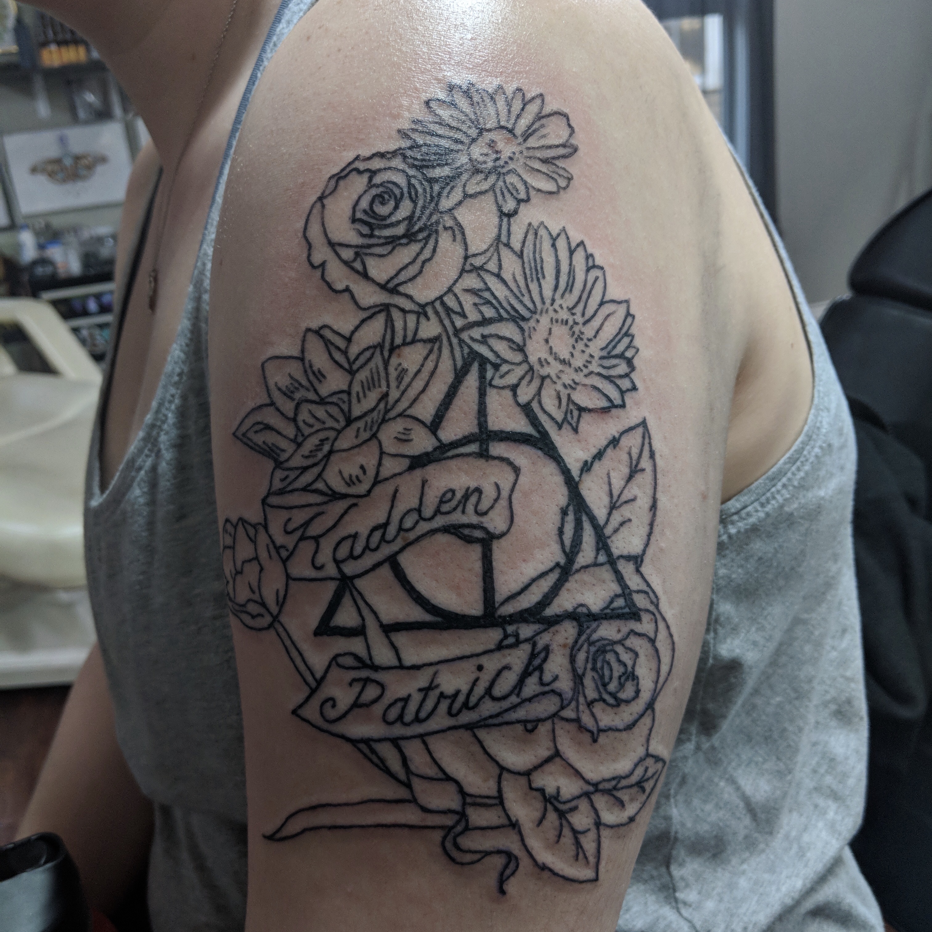 Image of an upper arm tattoo linework including a deathly hallows symbol, flowers and names