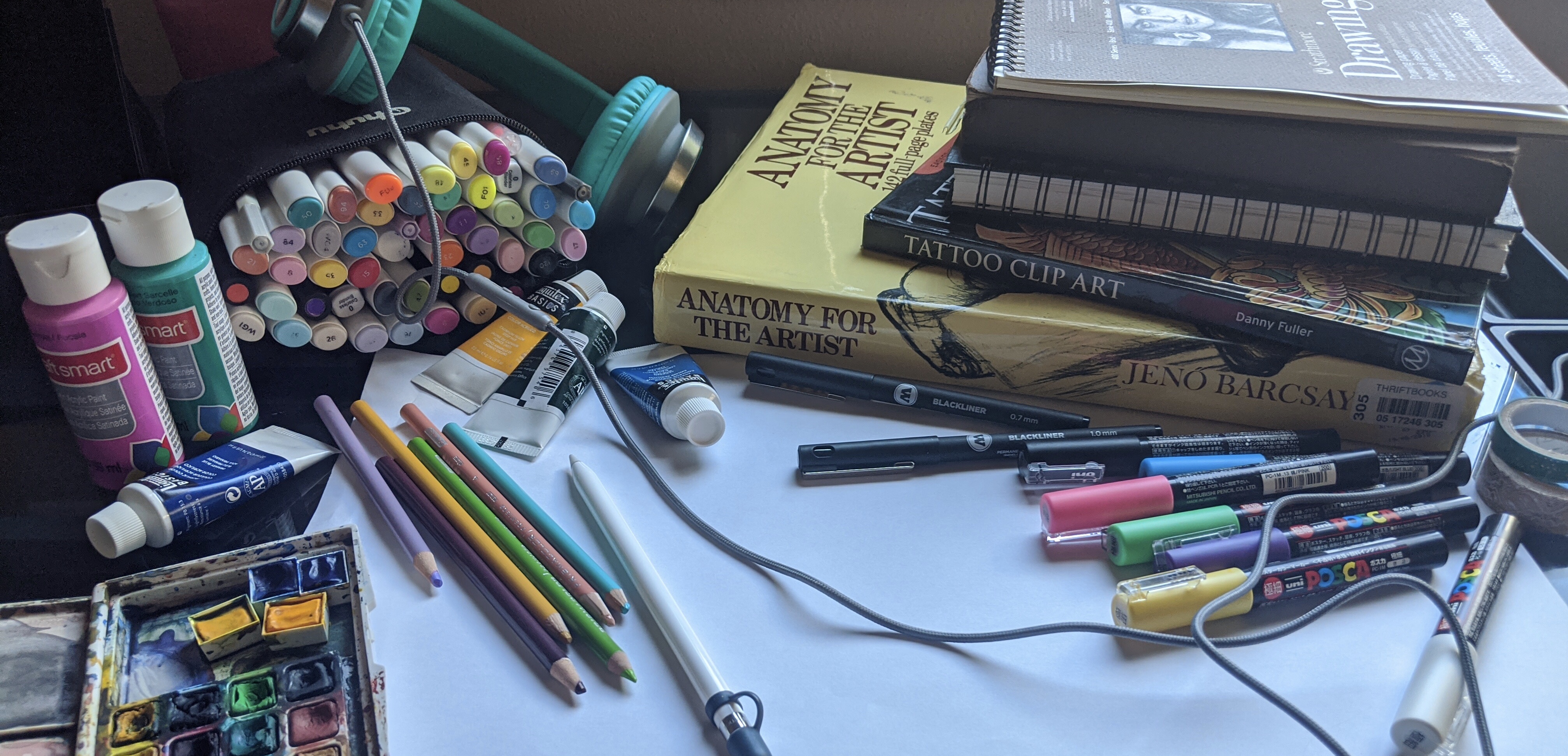 Image of art supplies and books on a desk. Acrylic paints, colored pencil, alcohol markers, watercolor and paint pens