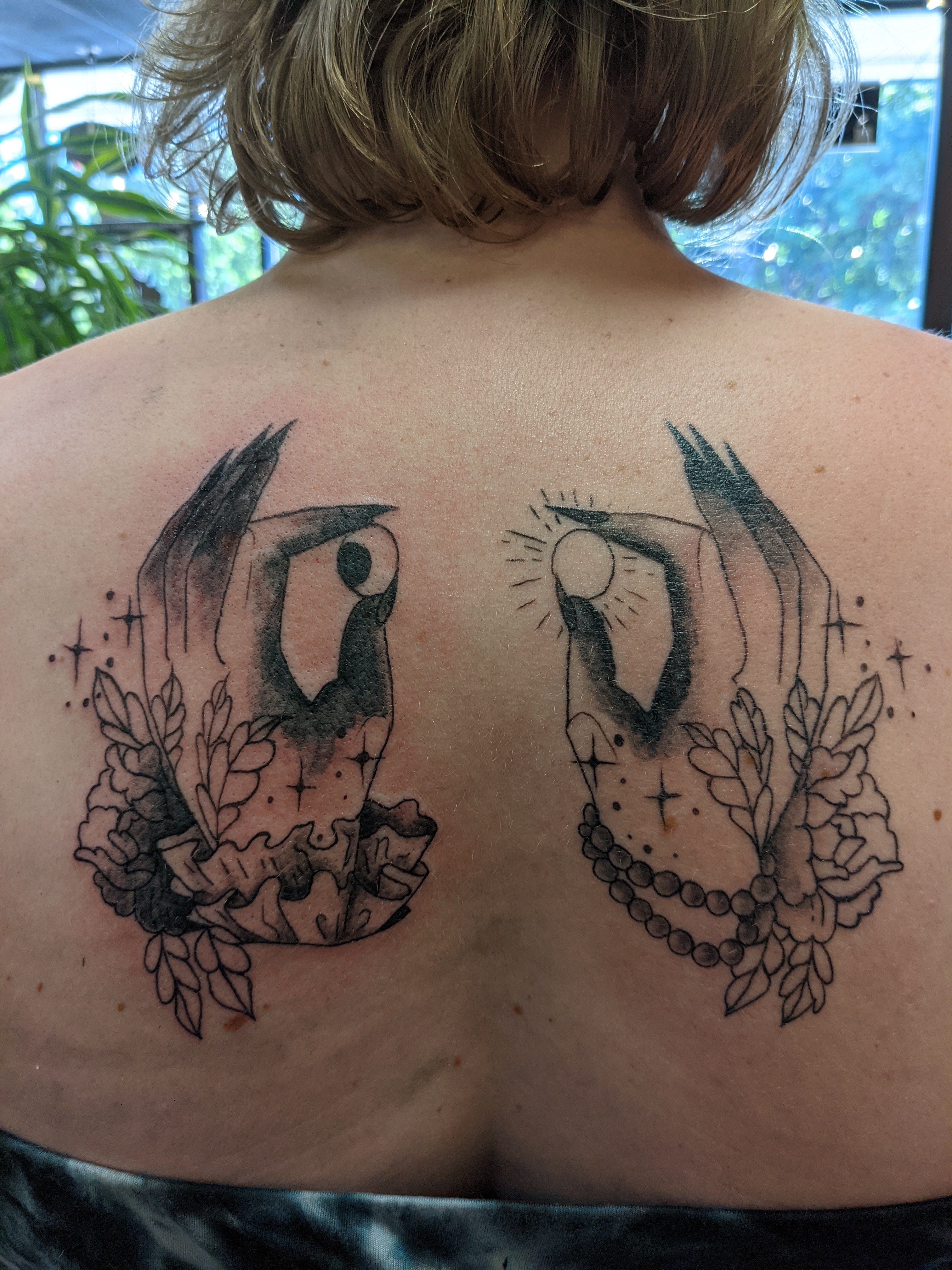 Image of two tattoos of hands on an upper back holding a moon and a sun with ruffled sleeves and jewelry